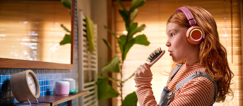 Malá dívka s růžovými svítícími sluchátkami Philips TAK4200 zpívající do hřebenu v koupelně